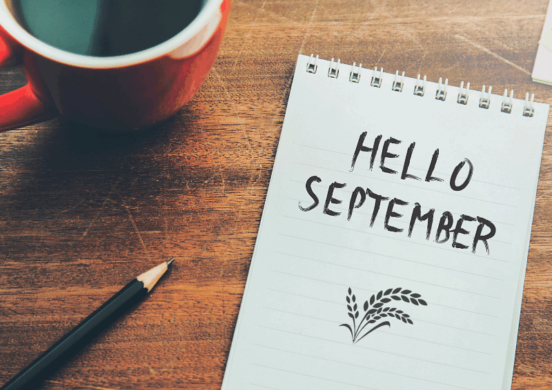 Coffee cup on a table with note pad and drawing pencil with "Hello September" drawn on the paper